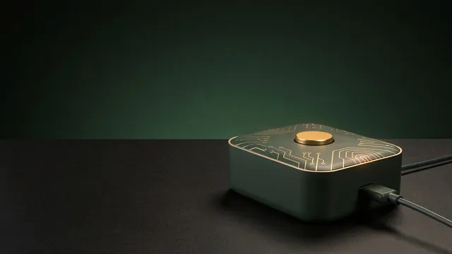 A small dark green box with one glowing gold button, plugged in on a desk.